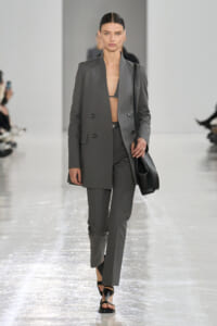 Model walking the runway in a gray double-breasted suit with blazer open at the chest, wearing a black bralette and carrying a black shoulder bag.