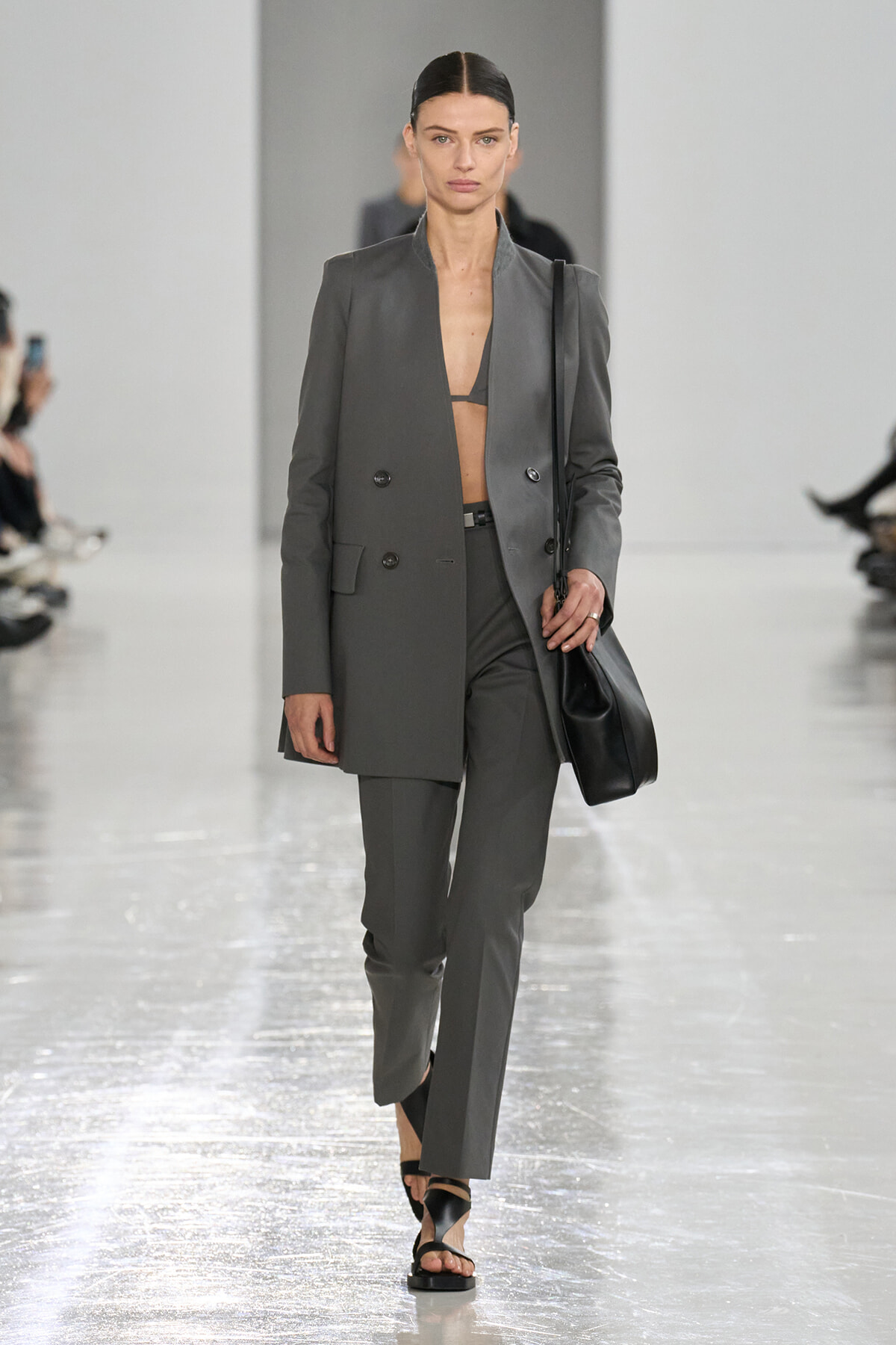 Model walking the runway in a gray double-breasted suit with blazer open at the chest, wearing a black bralette and carrying a black shoulder bag.