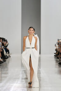 Model in a white halter gown with a black belt walks the glossy runway; photographers line the catwalk.