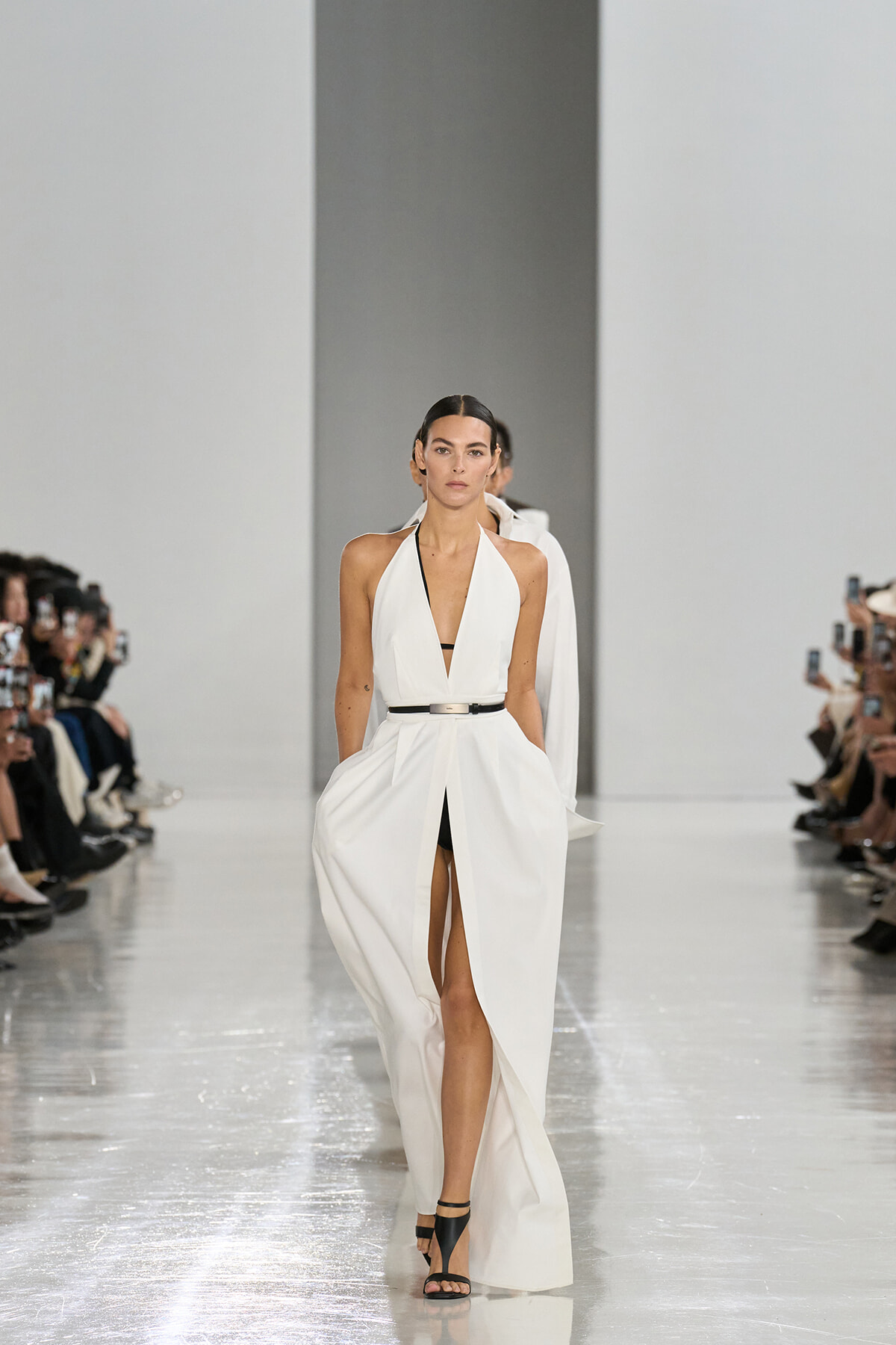 Model in a white halter gown with a black belt walks the glossy runway; photographers line the catwalk.