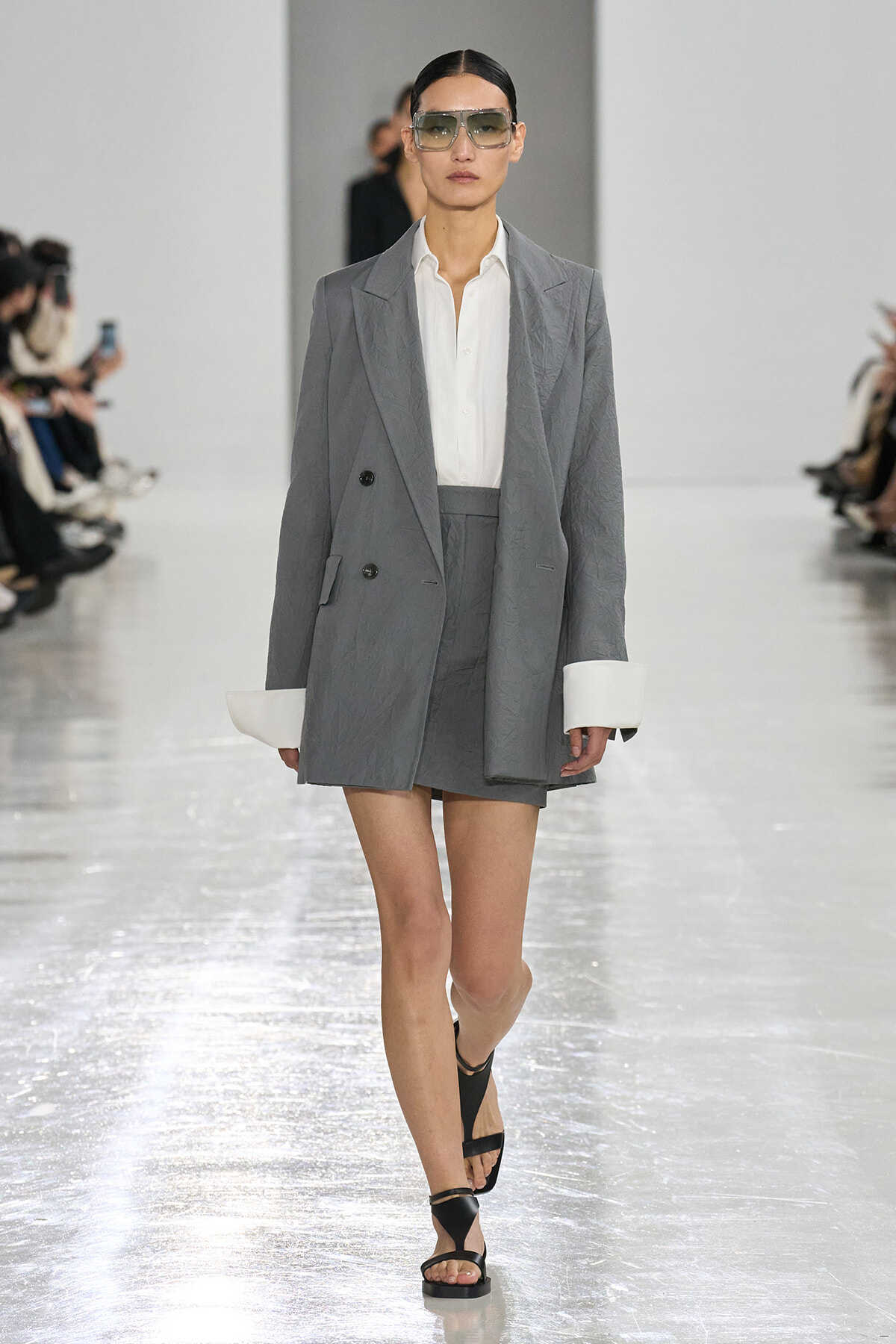 Model walking the runway in an oversized gray suit with a white shirt and matching shorts, accessorized with large sunglasses and black sandals.