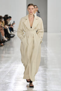 Model walking a fashion runway in a cream trench coat with wide lapels and a belt, hands in pockets.