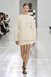 Model walks a minimalist cream-toned outfit down a runway with a loose top and short skirt, black sandals visible.