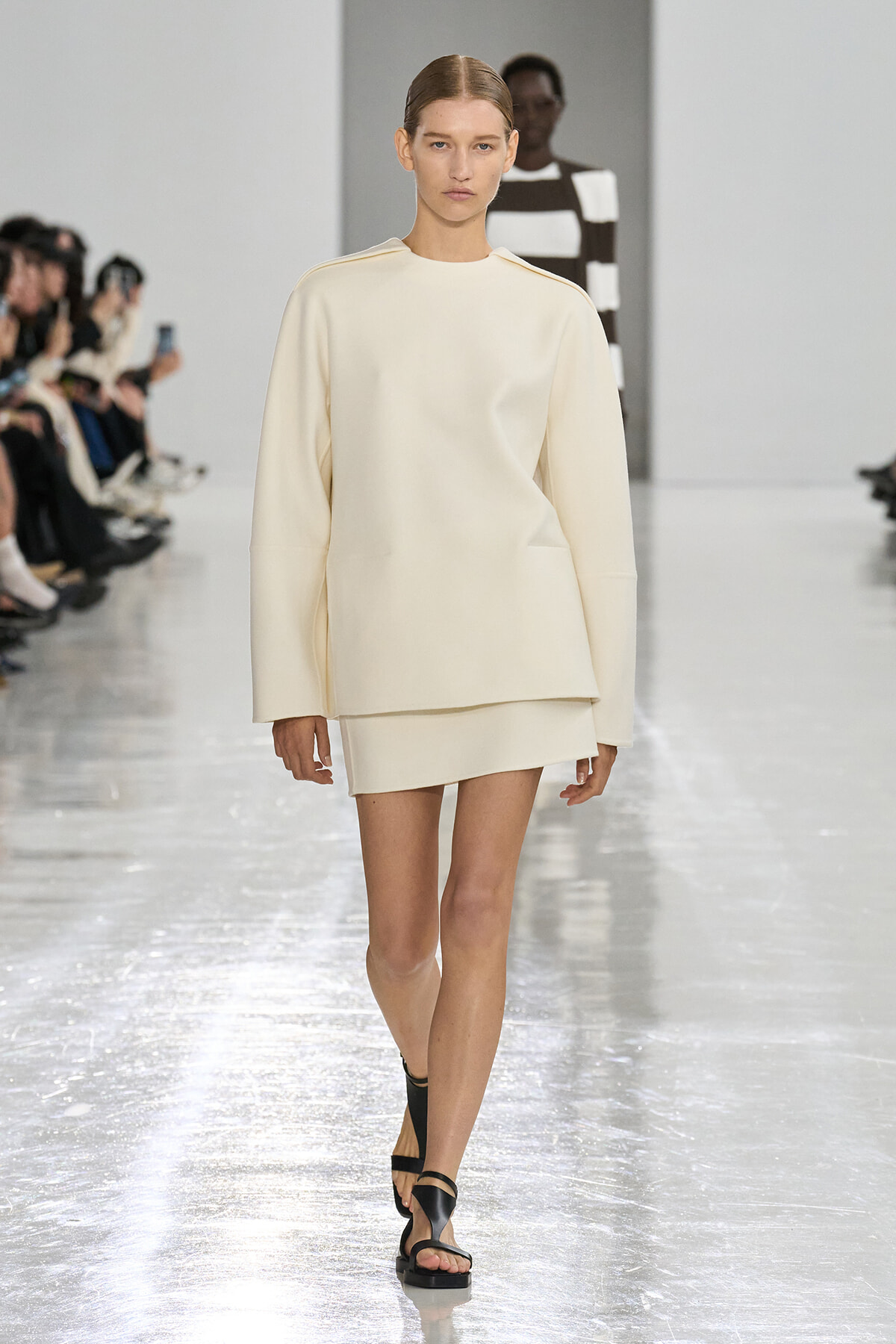 Model walks a minimalist cream-toned outfit down a runway with a loose top and short skirt, black sandals visible.