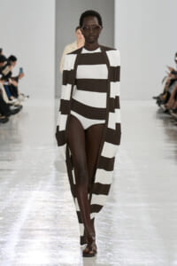 Runway model wearing a bold black-and-white striped coat over a matching striped bodysuit, with oversized sunglasses, walking the catwalk.