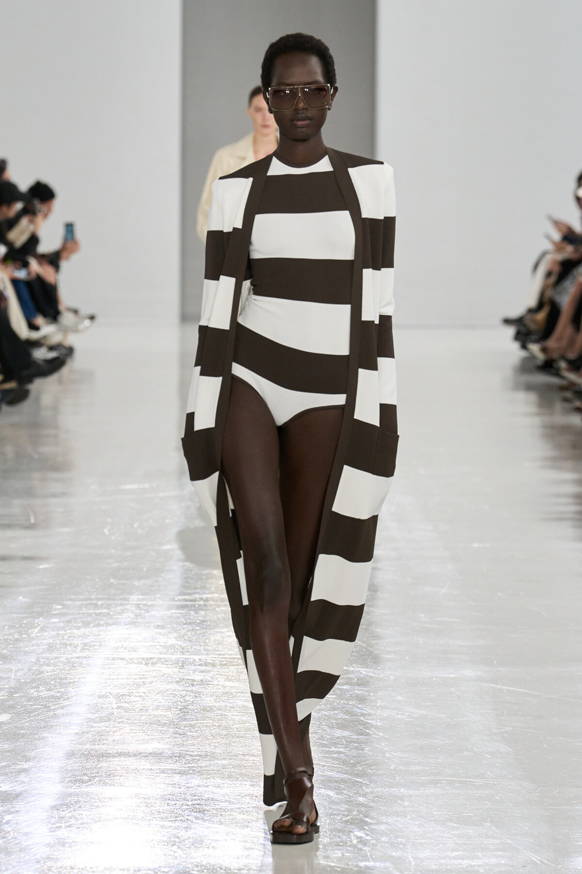 Runway model wearing a bold black-and-white striped coat over a matching striped bodysuit, with oversized sunglasses, walking the catwalk.