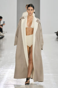 Model on a fashion runway wearing a long beige coat with white lapels over a cream bikini and black sandals.