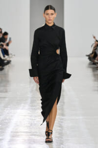 Model walks the runway in a black wrap dress with a waist cutout and high slit, paired with black sandals.