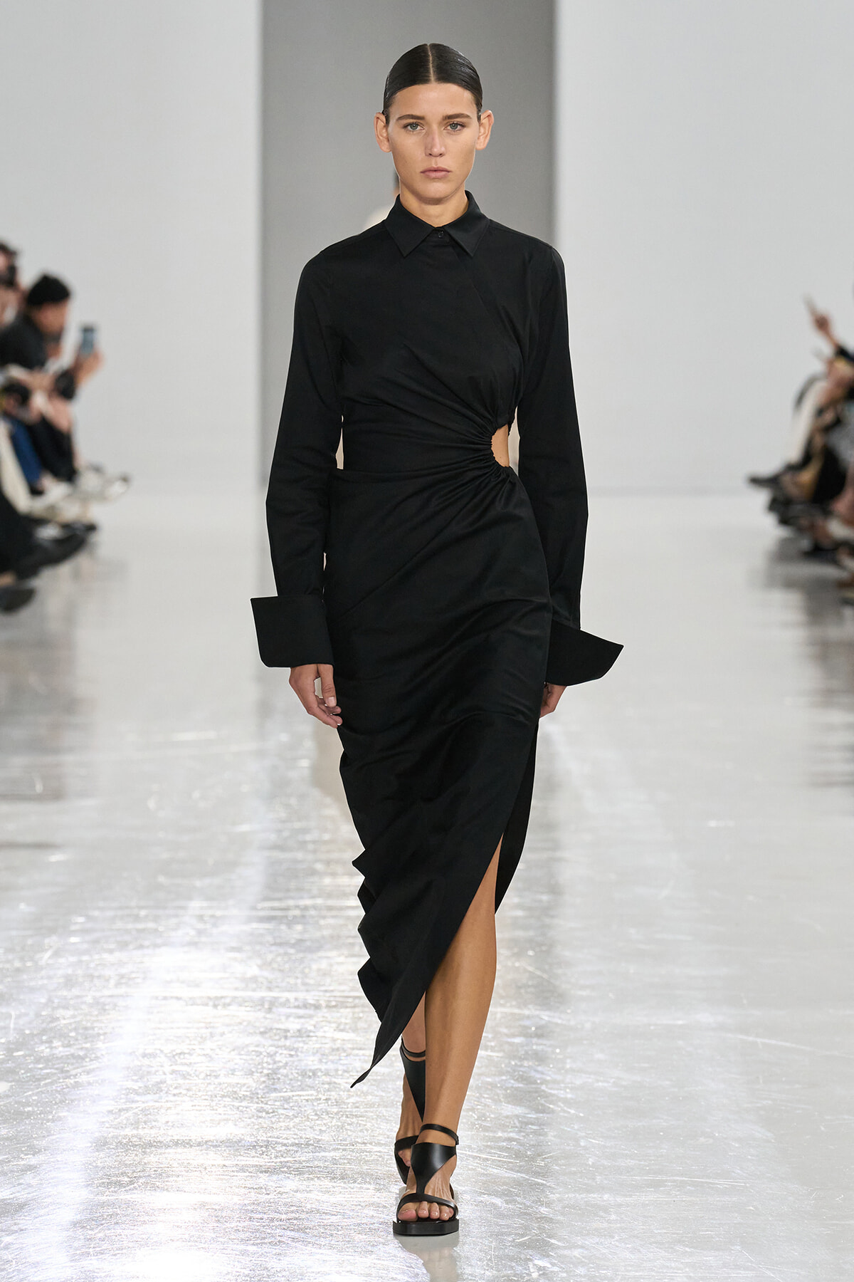 Model walks the runway in a black wrap dress with a waist cutout and high slit, paired with black sandals.