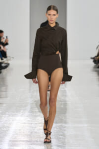 Model walking a glossy runway in a dark brown ensemble: twisted cropped blouse and high-waisted bottoms with strappy brown sandals.