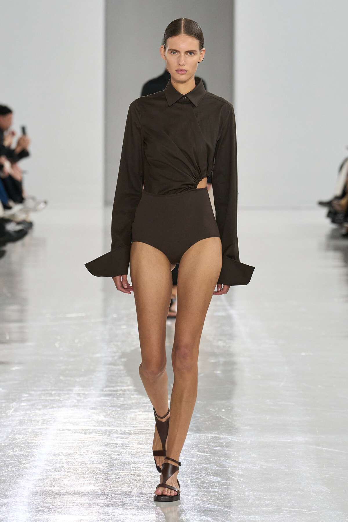 Model walking a glossy runway in a dark brown ensemble: twisted cropped blouse and high-waisted bottoms with strappy brown sandals.