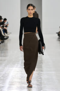 Model walks a fashion-show runway in a black ribbed cropped sweater and a brown wrap midi skirt, carrying a black handbag.