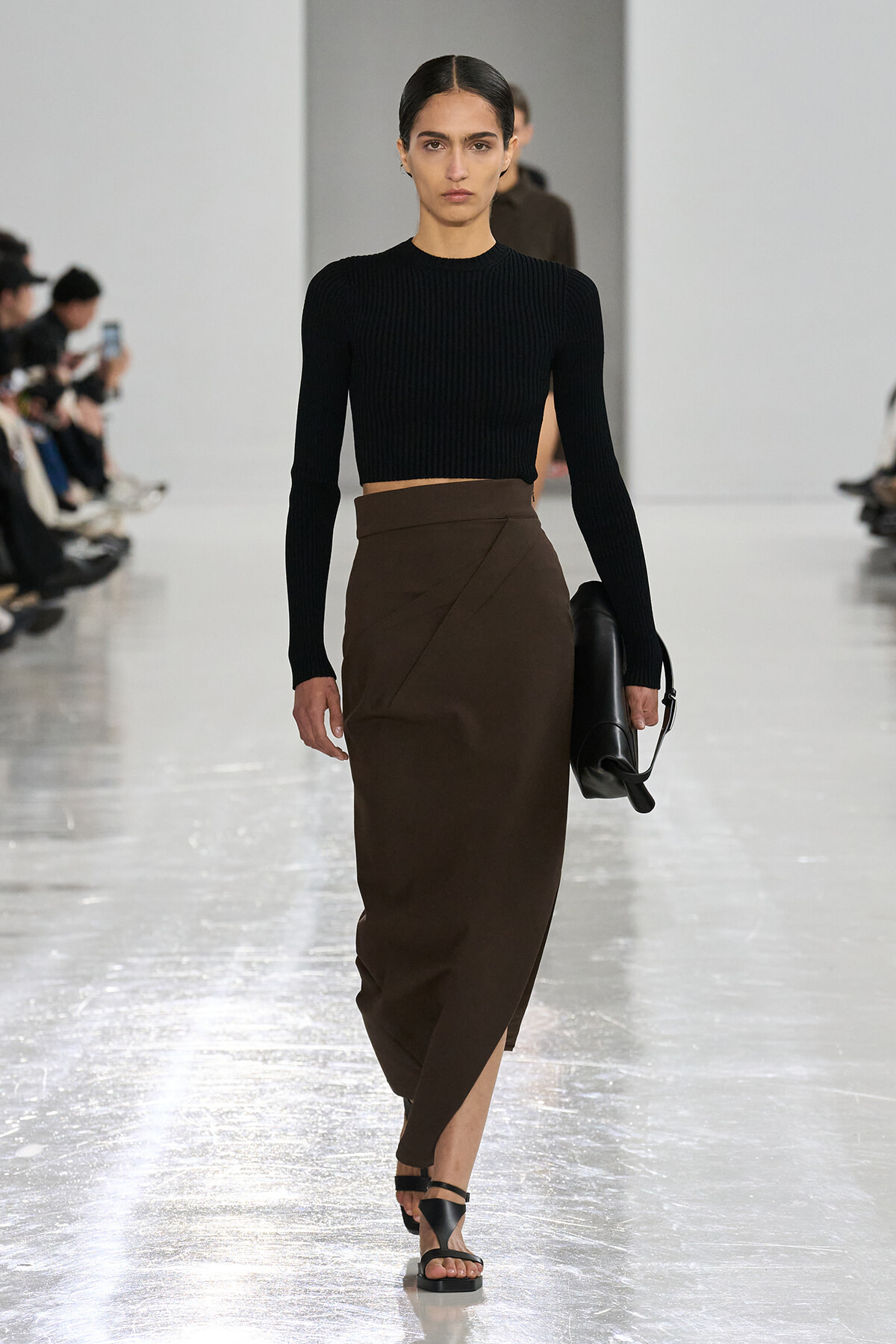 Model walks a fashion-show runway in a black ribbed cropped sweater and a brown wrap midi skirt, carrying a black handbag.