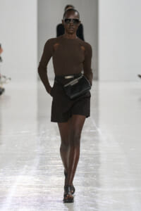 Model walking the runway in a brown ribbed knit top with shoulder cutouts, black shorts, a belt bag, and oversized sunglasses.
