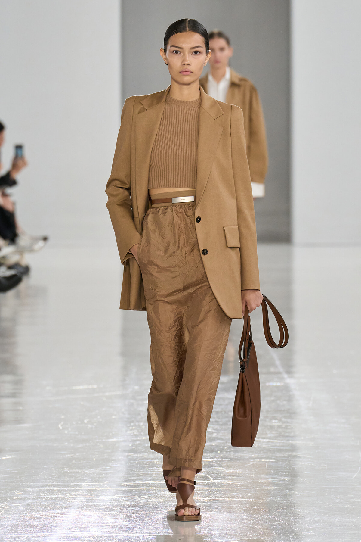 Model walks a fashion show wearing a camel blazer over a ribbed tan top and crinkled tan trousers, carrying a brown handbag.