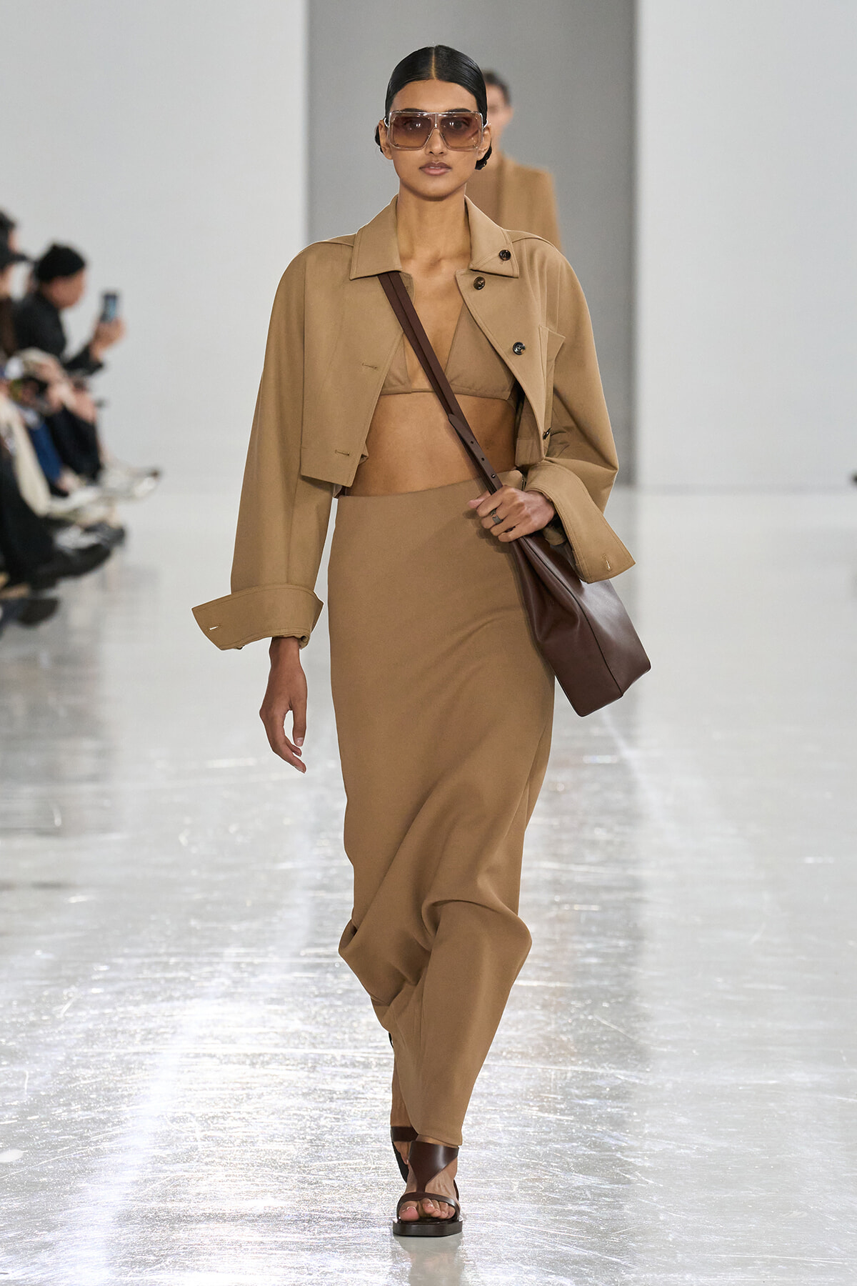 Model walks the runway in a camel cropped coat, bralette, and long skirt with a brown crossbody bag and oversized sunglasses.