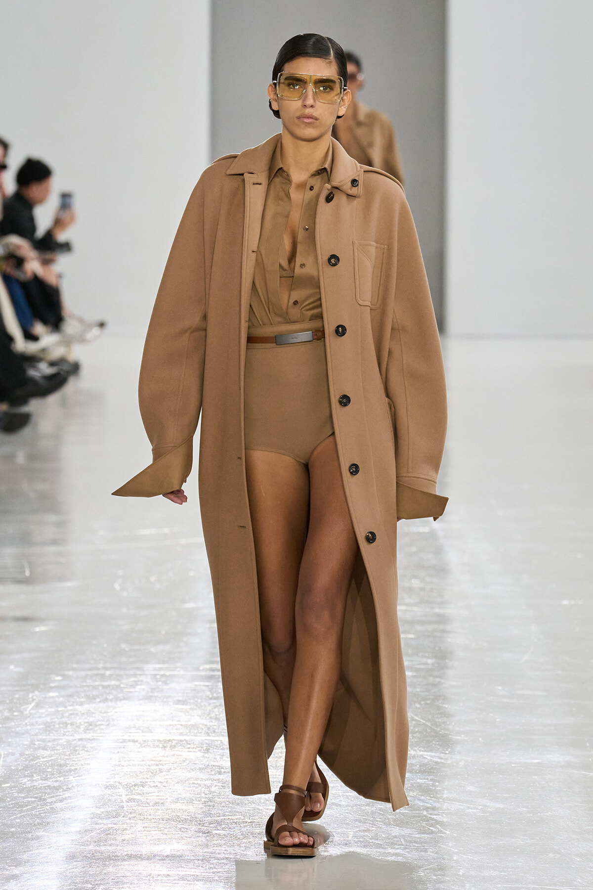 Runway model walks in a camel coat over a tan bodysuit with a brown belt and brown sandals, wearing oversized yellow-tinted sunglasses.
