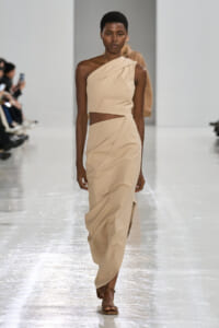 Model walks a beige one-shoulder crop top and matching wrap skirt on a glossy runway at a fashion show