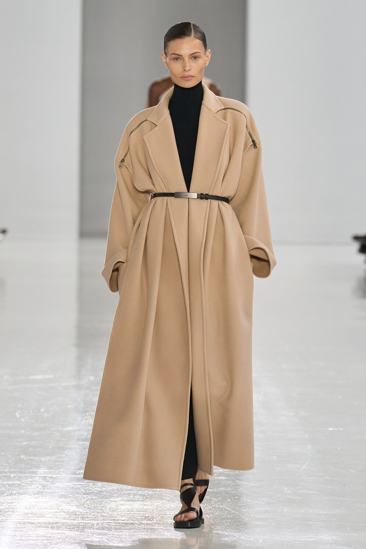 Model walks a runway in a long beige oversized coat belted at the waist over a black turtleneck, paired with black sandals.