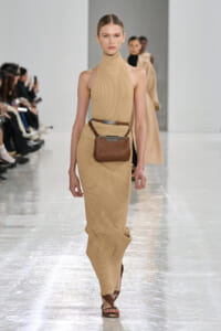 Model walks the runway in a beige ribbed sleeveless turtleneck dress with a brown belt bag and sandals on a glossy catwalk.
