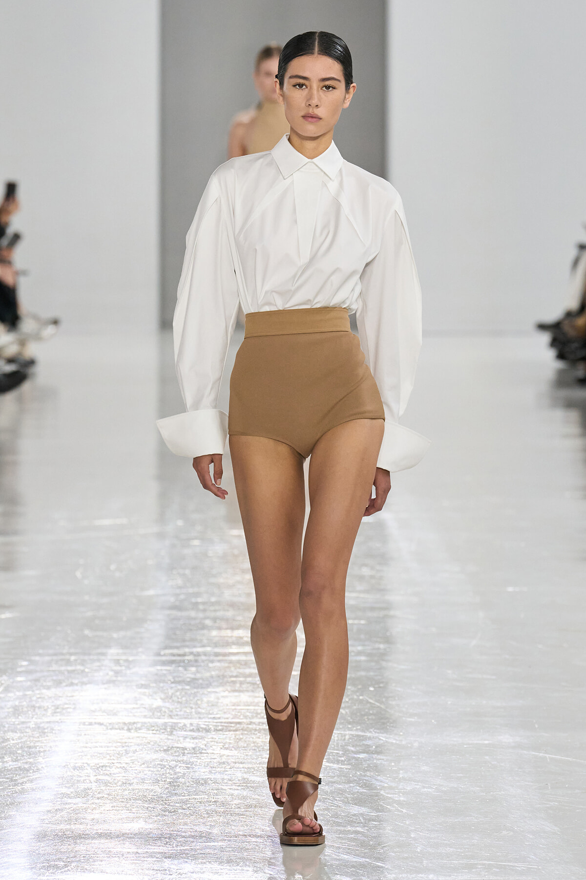 Model walking the runway in a white oversized shirt tucked into tan high-waisted shorts with brown sandals.