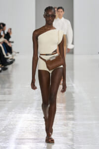 Runway model wearing a cream one-shoulder knit top with matching high-waisted bottoms and a brown belt bag.