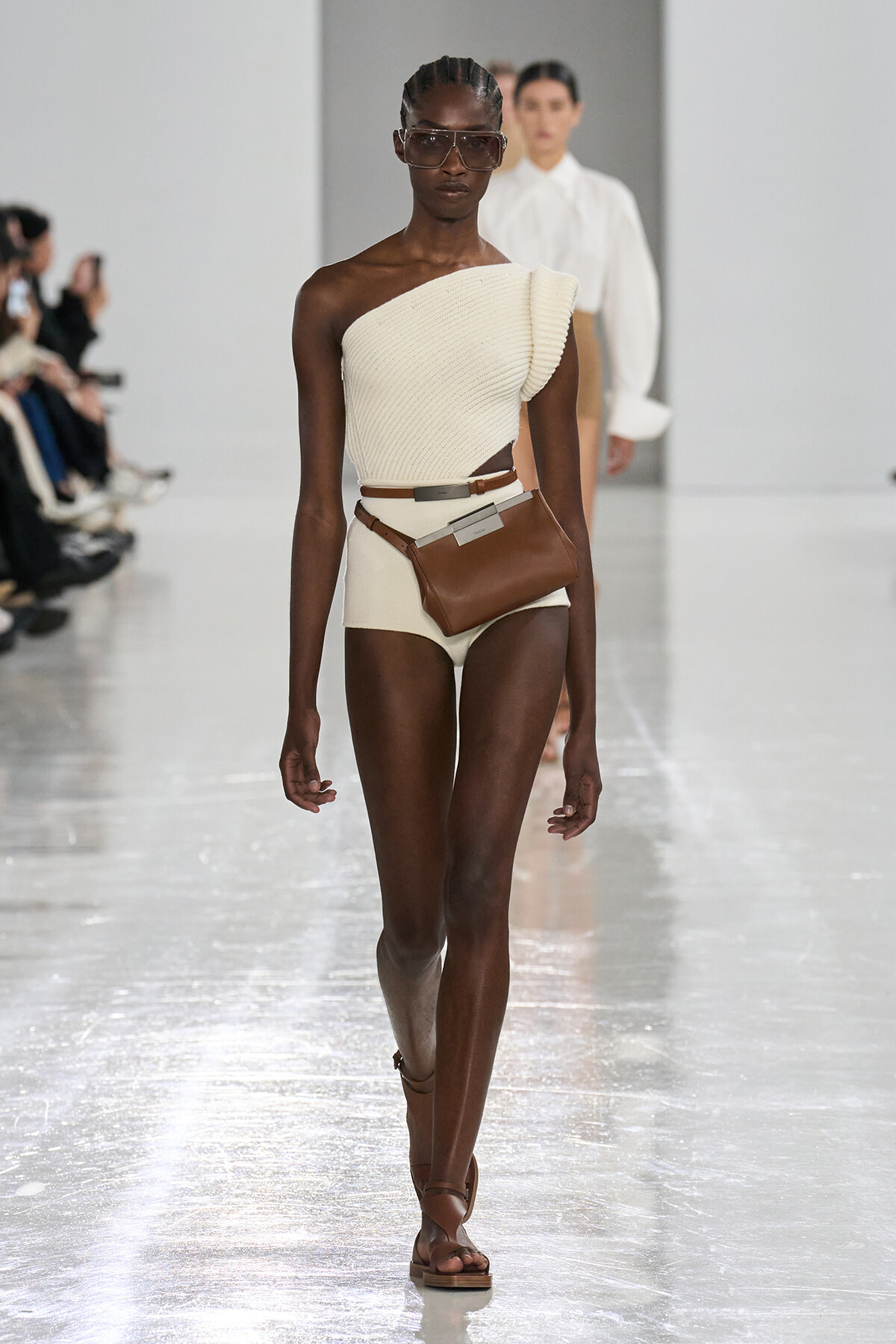 Runway model wearing a cream one-shoulder knit top with matching high-waisted bottoms and a brown belt bag.