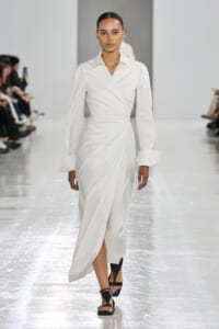 Model walks a runway in a white wrap dress with long sleeves and wide cuffs, paired with black strappy sandals.