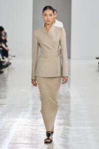 Model walking the runway in a beige wrap blazer with a tied waist and a ribbed beige midi skirt, paired with black sandals.