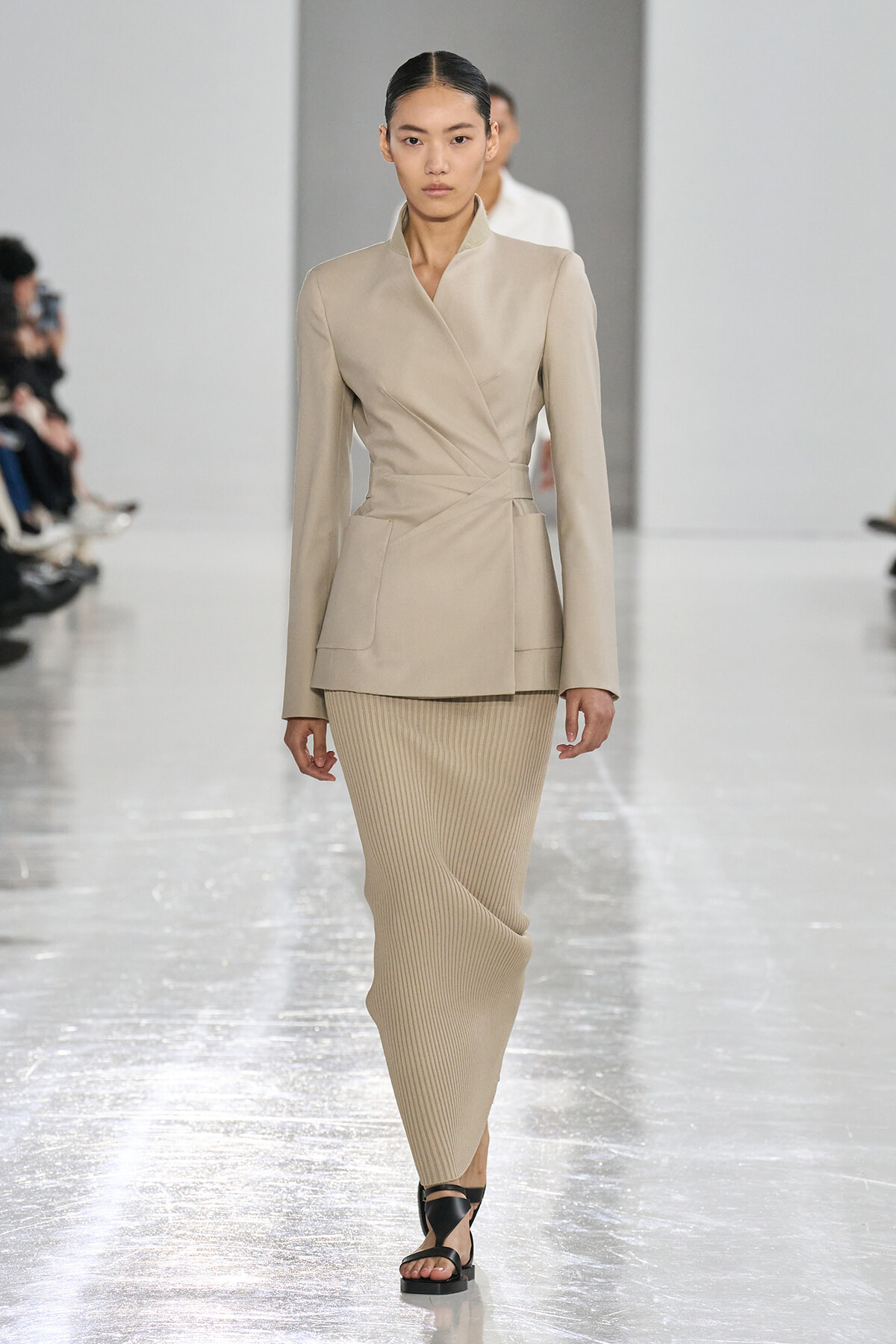 Model walking the runway in a beige wrap blazer with a tied waist and a ribbed beige midi skirt, paired with black sandals.