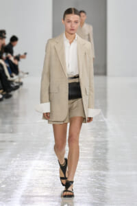 Runway model wearing a beige blazer over a white shirt with matching shorts and black sandals, walking on a glossy catwalk.