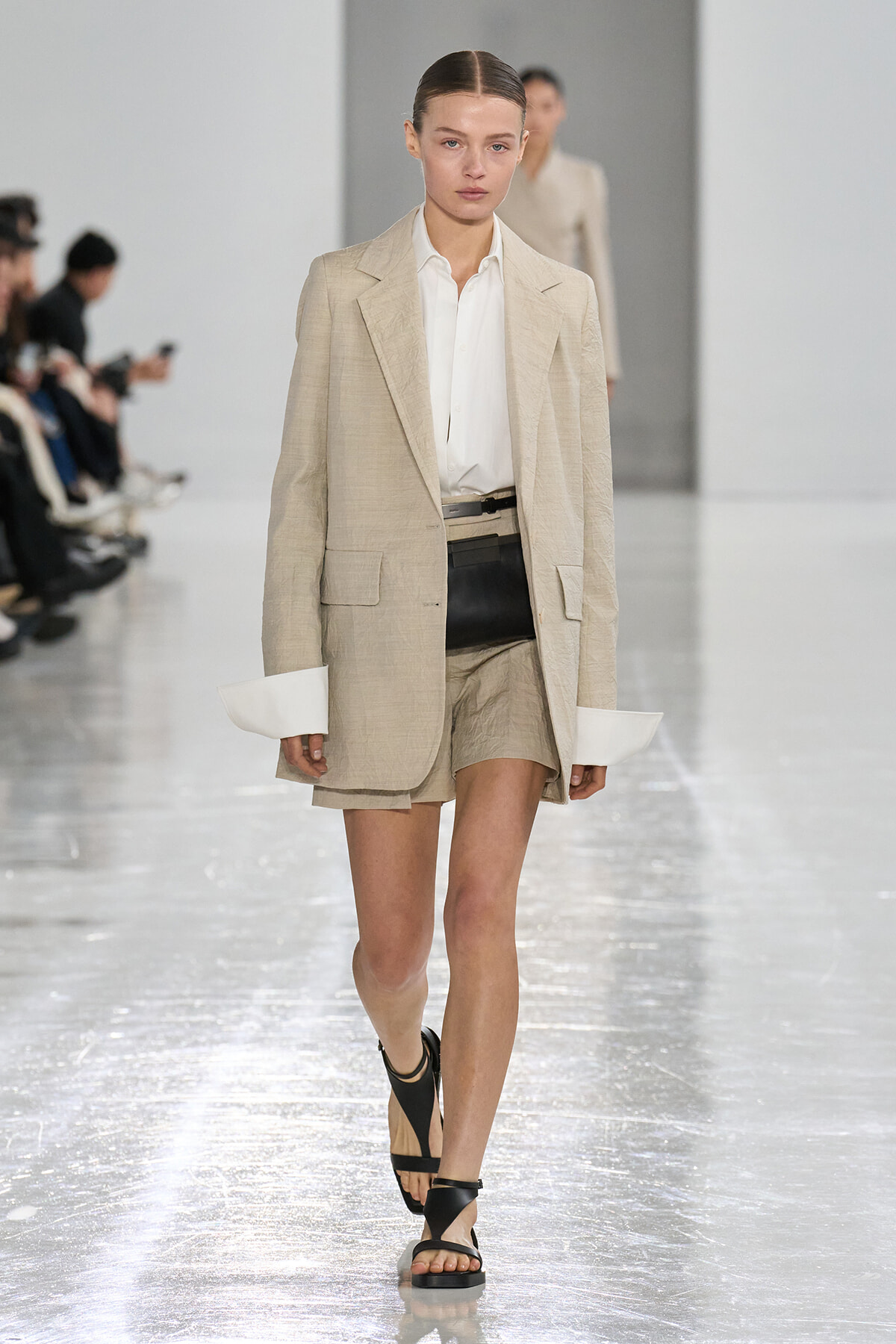 Runway model wearing a beige blazer over a white shirt with matching shorts and black sandals, walking on a glossy catwalk.