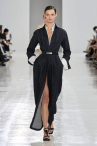 Model walks a dark denim gown with white cuffs, belted waist, and a front slit revealing her leg on a fashion runway.