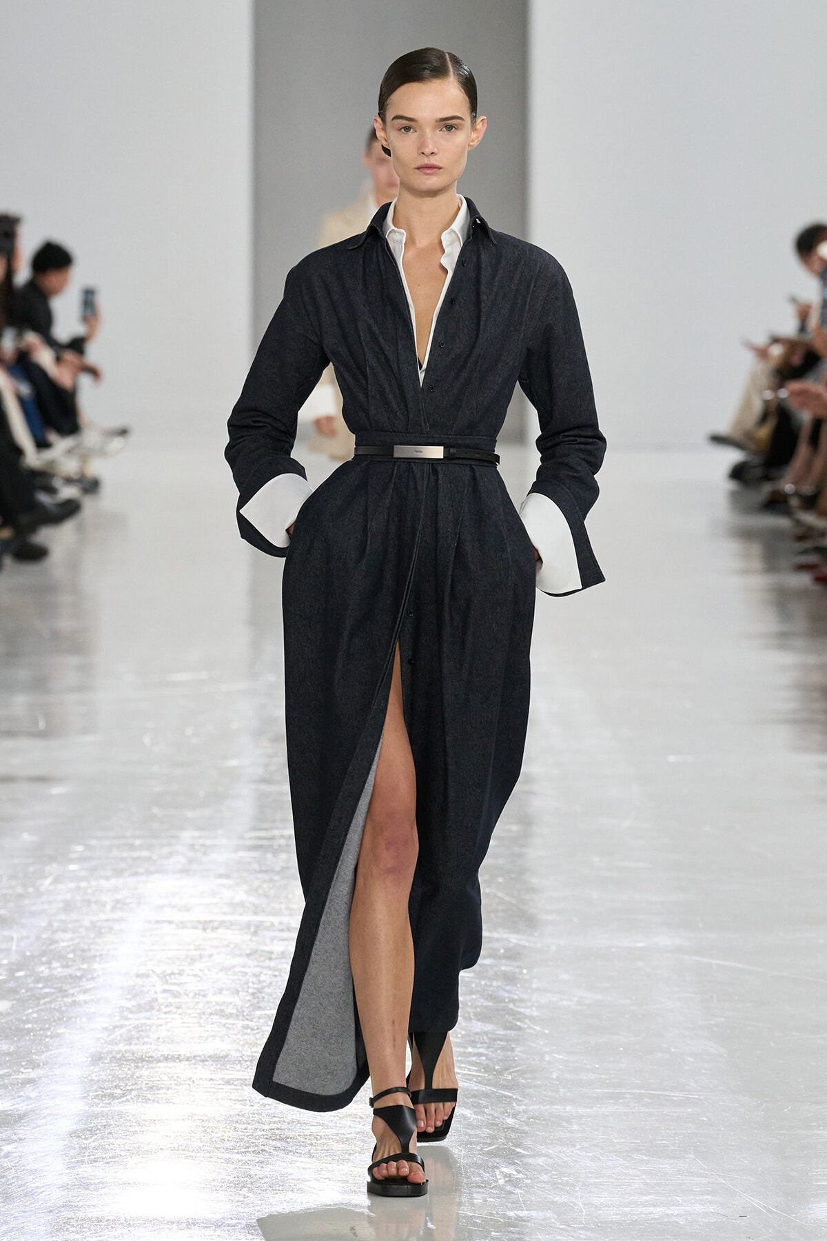 Model walks a dark denim gown with white cuffs, belted waist, and a front slit revealing her leg on a fashion runway.