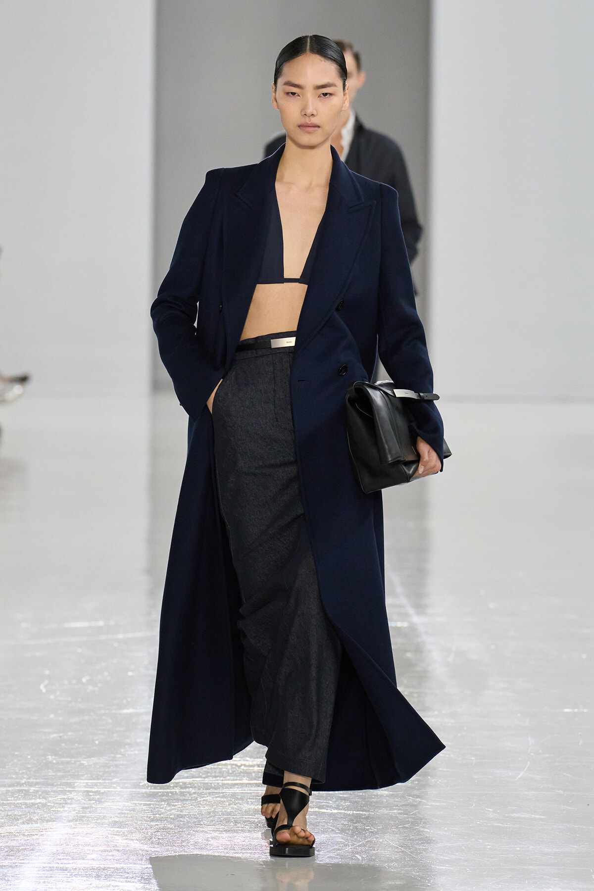 Model walks runway in a navy long coat opened over a navy bralette and gray wide-leg trousers, carrying a black clutch.