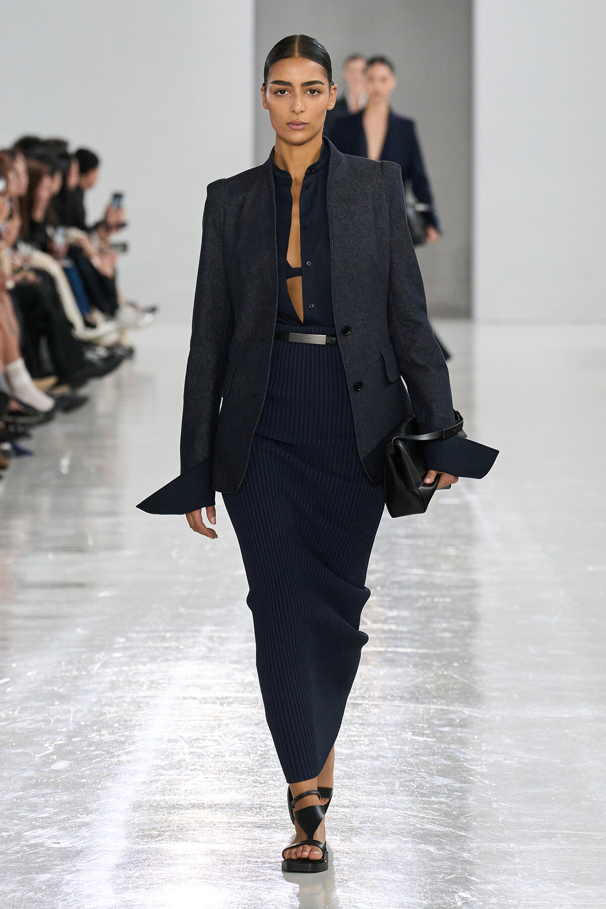 Female model walks a fashion show in a navy blazer and ribbed midi skirt, holding a black clutch and wearing black sandals.