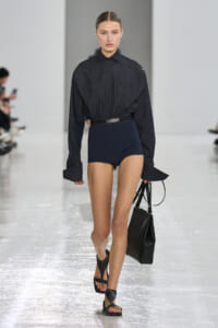 Model walking a runway wearing a dark denim shirt tucked into navy high-waisted shorts with a black belt, carrying a black handbag and wearing black sandals.