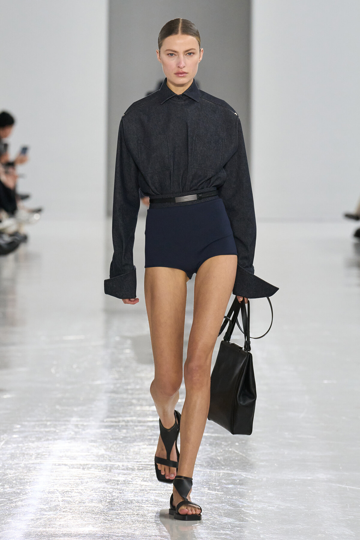 Model walking a runway wearing a dark denim shirt tucked into navy high-waisted shorts with a black belt, carrying a black handbag and wearing black sandals.