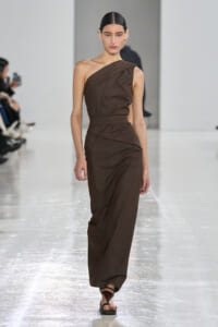 Female model walks a brown one-shoulder wrap dress with a belt, paired with flat sandals, on a runway.