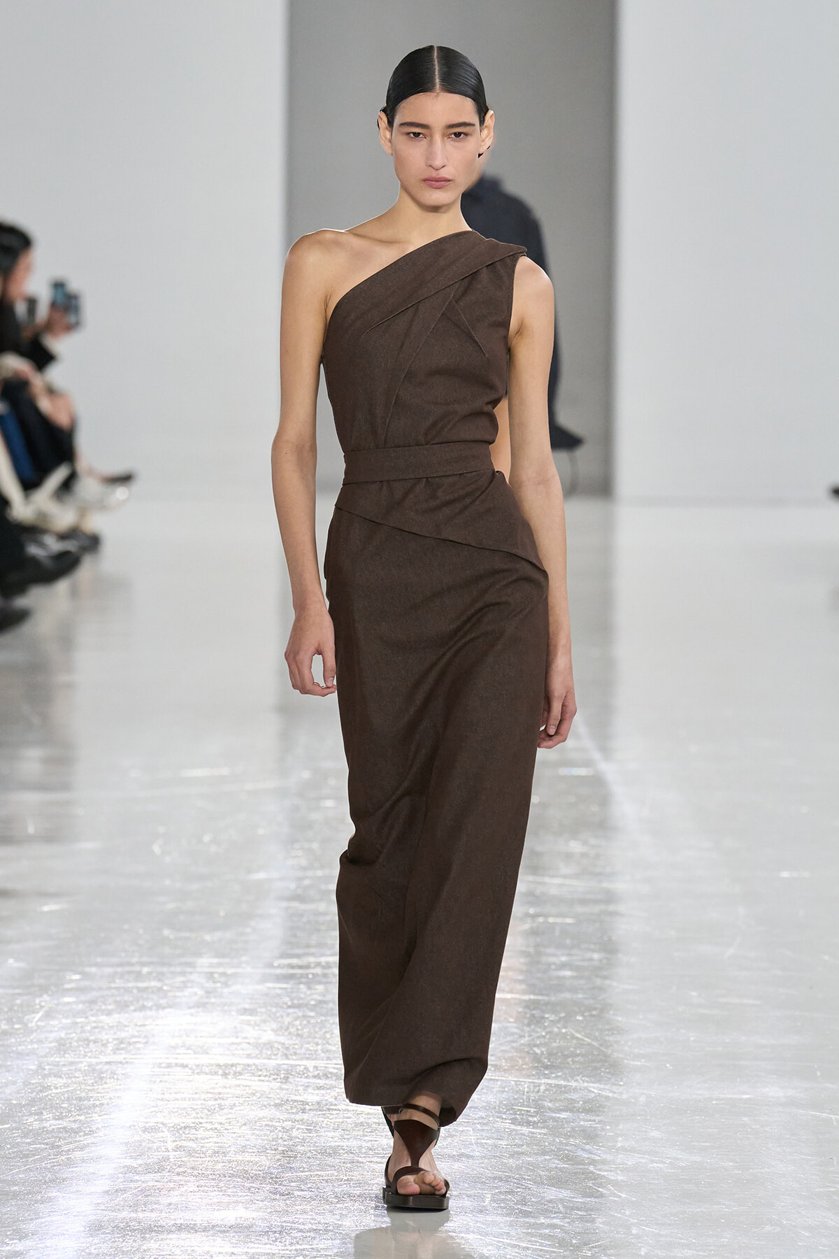 Female model walks a brown one-shoulder wrap dress with a belt, paired with flat sandals, on a runway.
