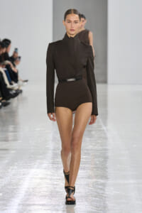 Model on a runway wearing a black high-neck blouse tucked into brown high-waisted shorts with a belt and black sandals.