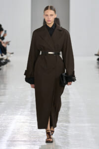Model walking the runway in a dark brown belted trench coat over a black shirt, holding a black clutch with brown sandals visible at the hem.