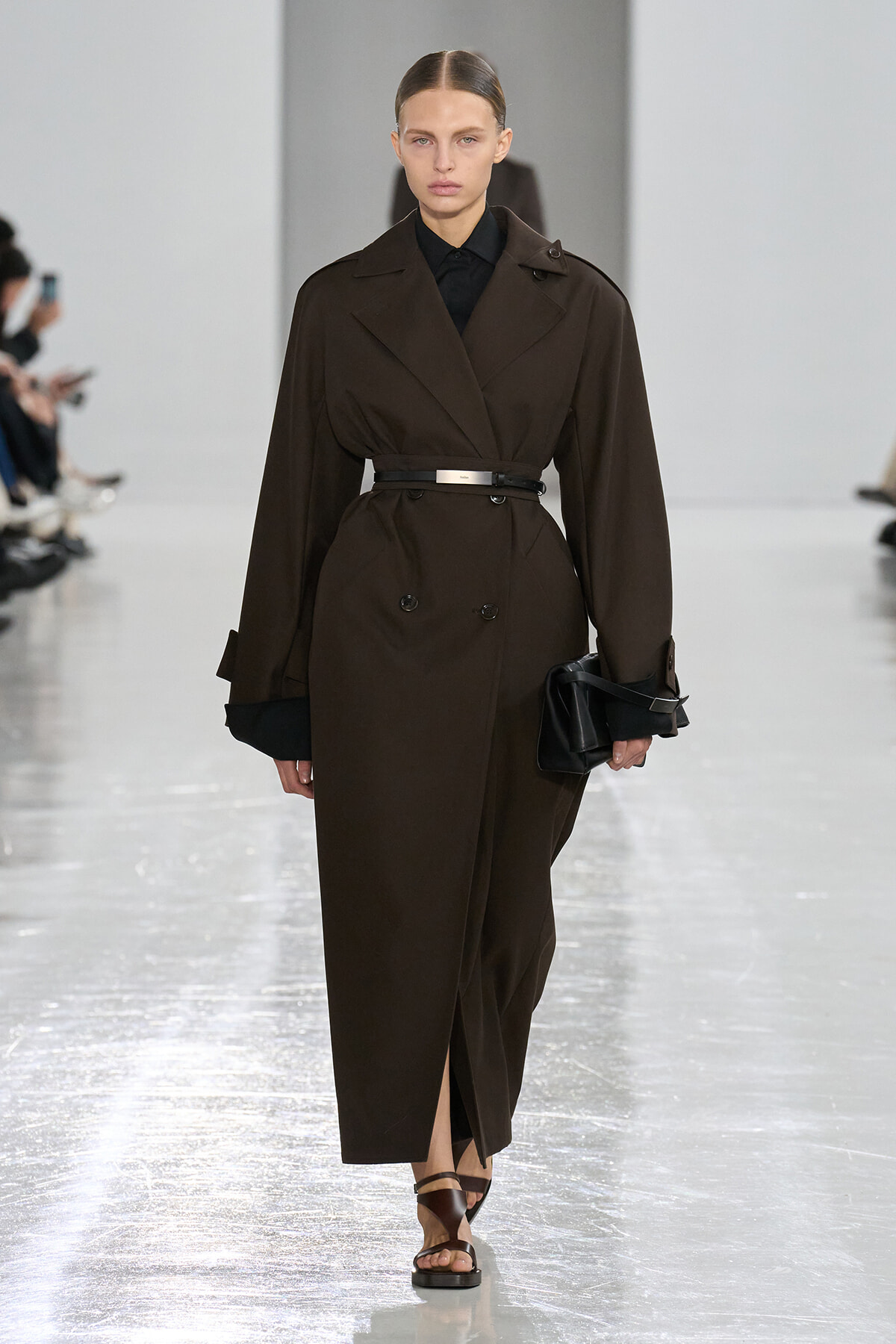 Model walking the runway in a dark brown belted trench coat over a black shirt, holding a black clutch with brown sandals visible at the hem.