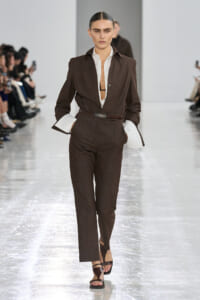 Model walks the runway in a dark brown tailored suit with white shirt cuffs peeking out, belt at the waist, and strappy sandals.