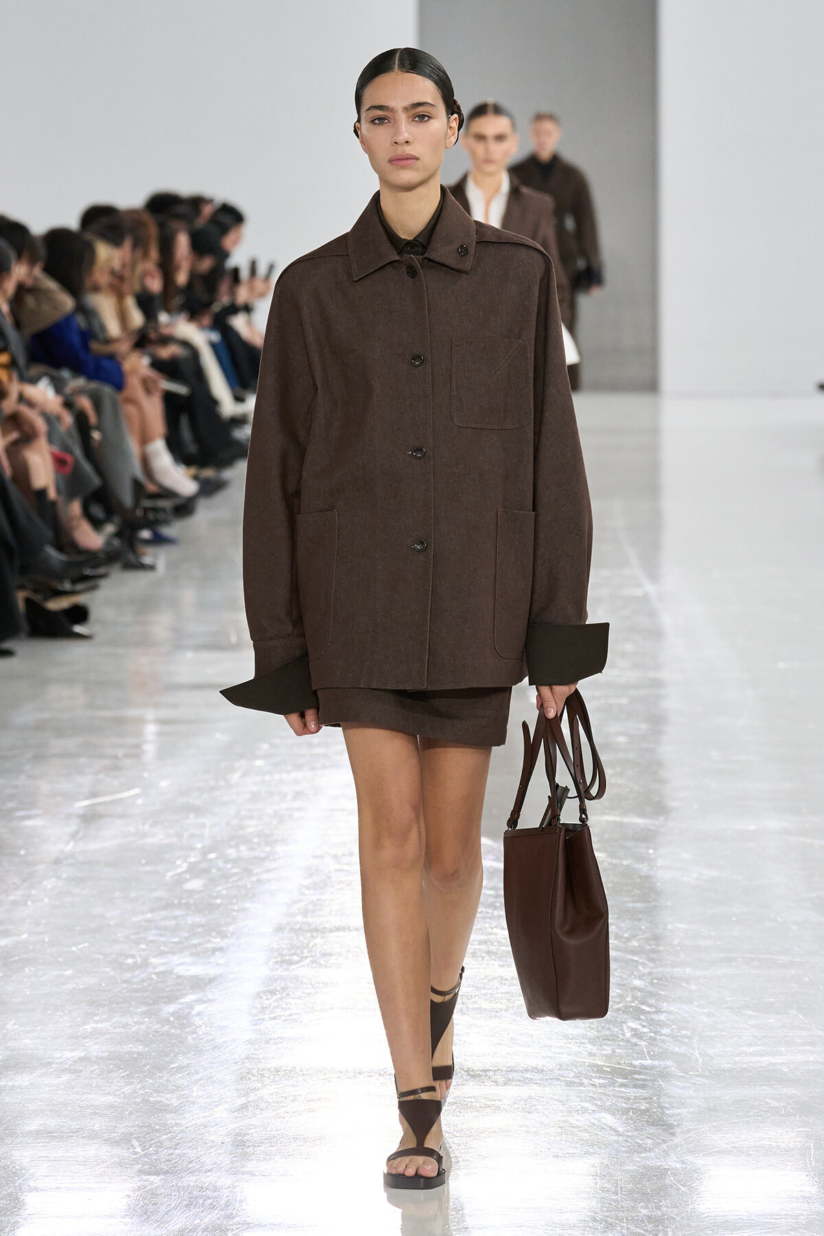 Model walking a fashion runway in a oversized brown coat with large pockets, matching shorts, and a brown tote bag.
