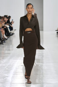 Model on a runway wearing a dark brown cropped blouse and a high-waisted ribbed brown skirt with a belt, sandals visible at the feet.