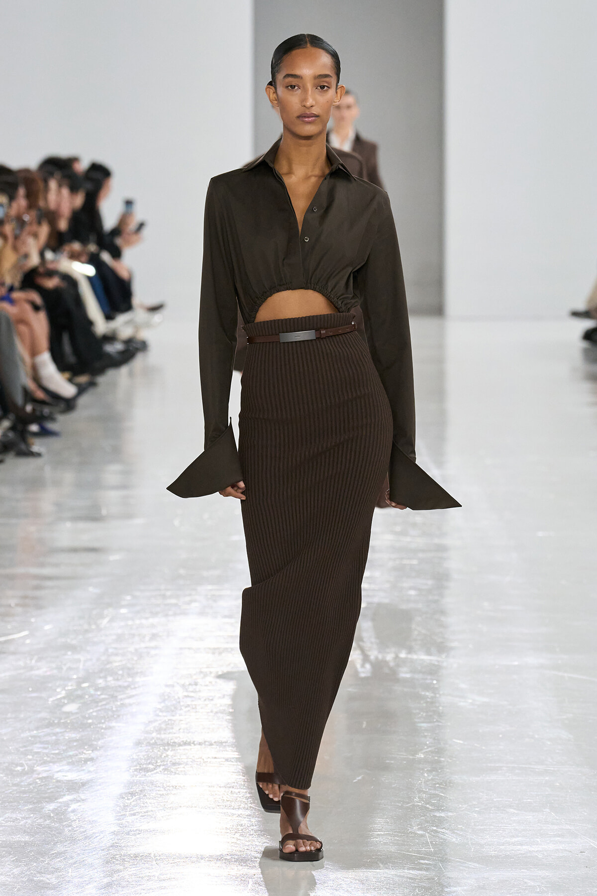 Model on a runway wearing a dark brown cropped blouse and a high-waisted ribbed brown skirt with a belt, sandals visible at the feet.
