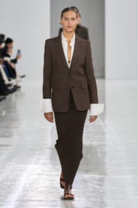 Model walking a fashion runway in a brown blazer over a white shirt and a long dark ribbed skirt, paired with brown sandals.