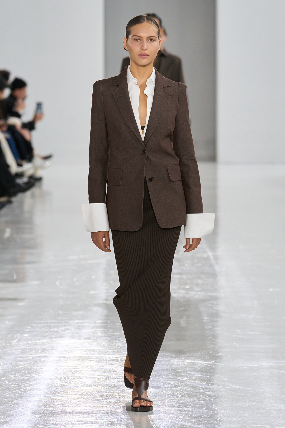 Model walking a fashion runway in a brown blazer over a white shirt and a long dark ribbed skirt, paired with brown sandals.