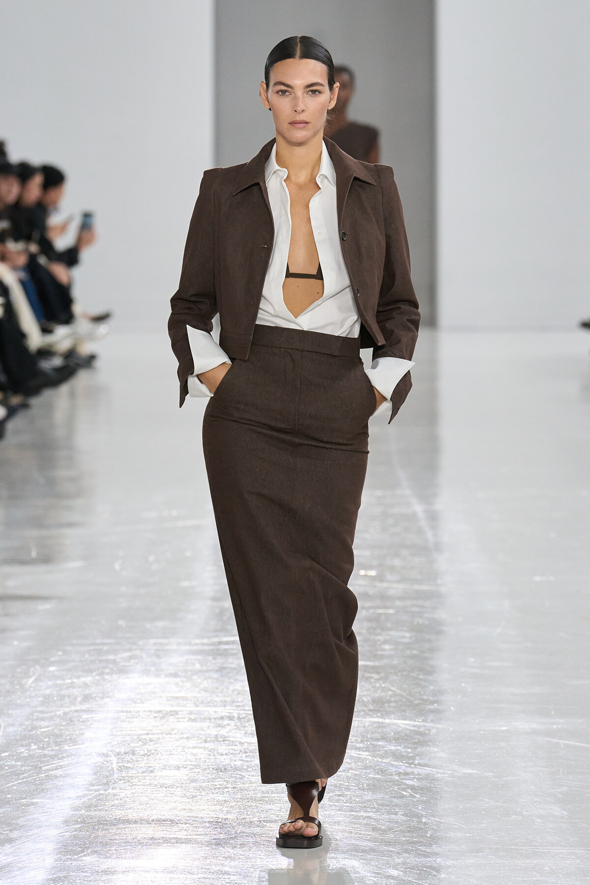 Model walks a fashion runway in a brown tailored blazer and matching high-waisted skirt with a white shirt, hands in pockets.